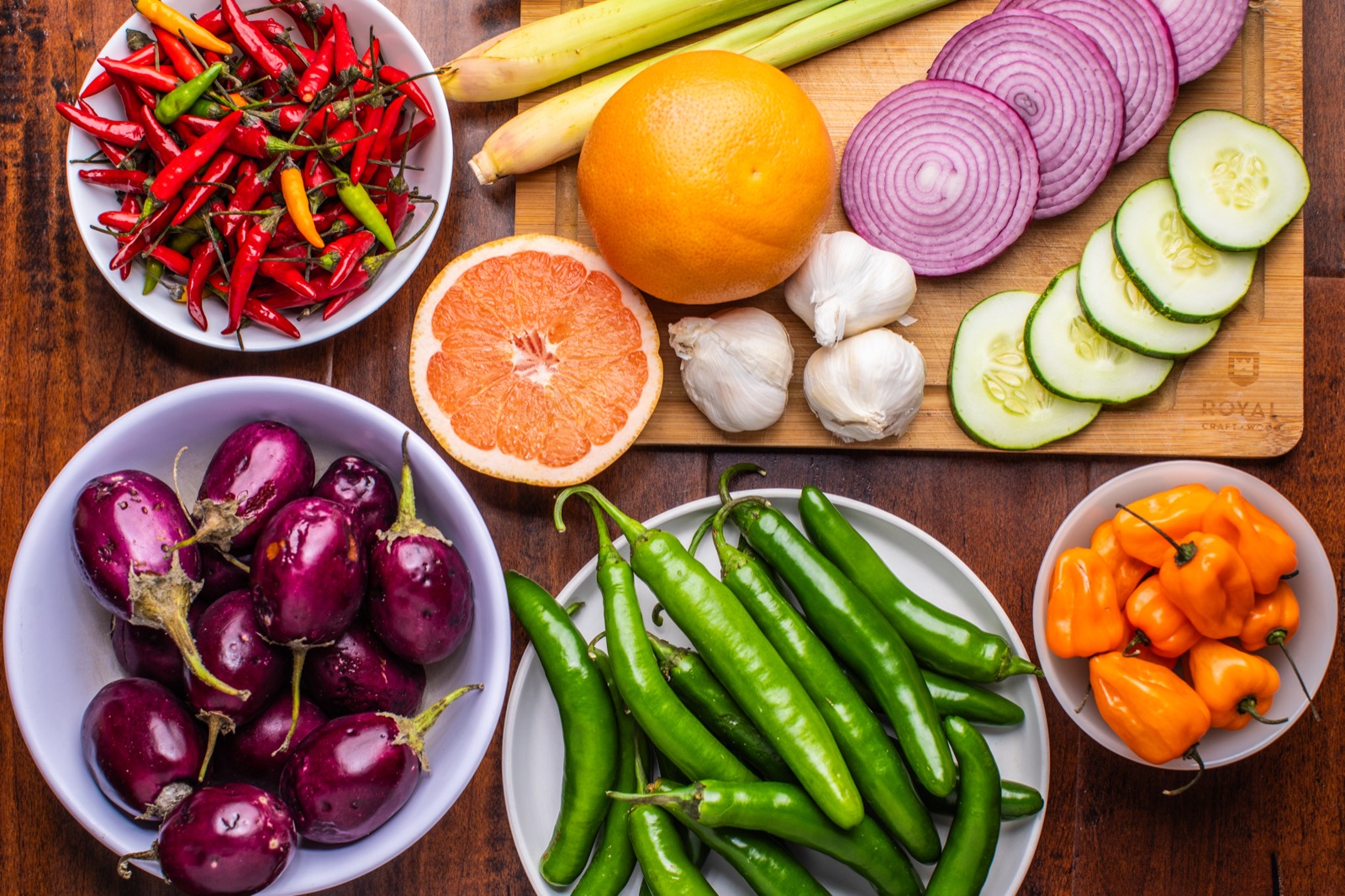 Fresh Asian specialty produce — chilies, Thai eggplant, grapefruit, lemongrass, garlic, red onion, and cucumber laid out on a wooden cutting board