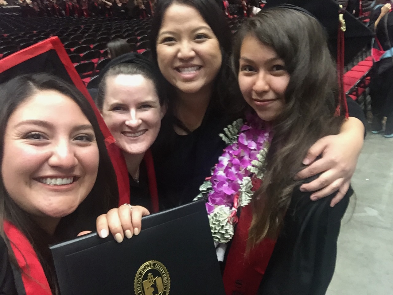 Michelle Xiong celebrating with three graduating Liberal Studies students in cap and gown at an SDSU commencement ceremony, one holding her diploma and another wearing a flower lei.